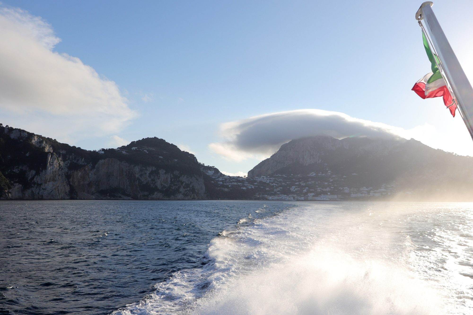 A catamaran sailing off Capri with sails up and the coastline behind